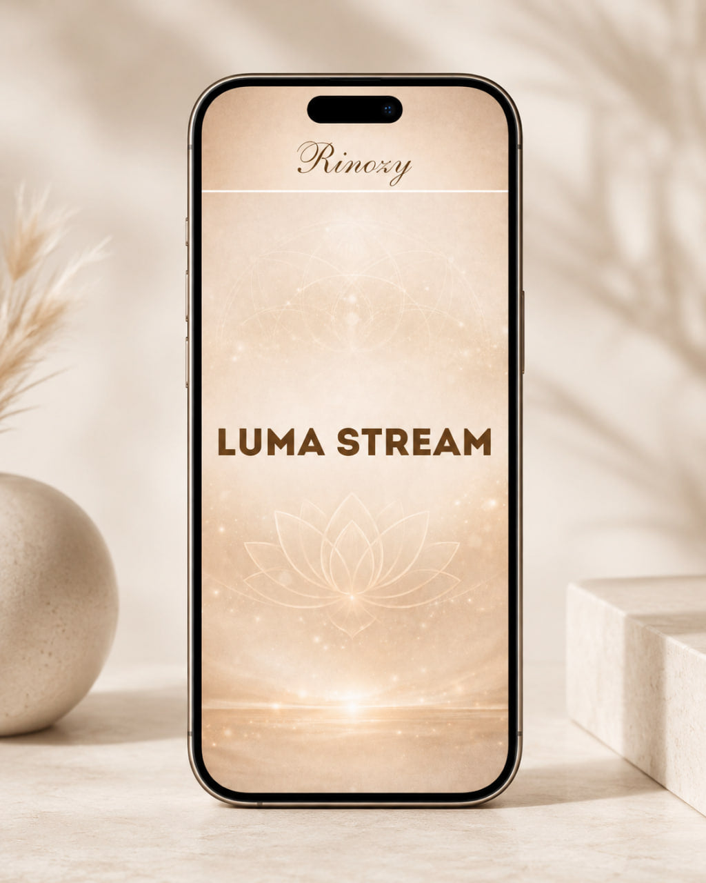 A smartphone centered on a light beige surface with soft decorative props in the background. The phone screen displays the text 'Rinozy' at the top and 'LUMA STREAM' in large brown letters over a warm cream and gold abstract design.